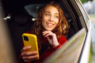 Genç bir kadın arabanın arka koltuğunda otururken akıllı telefon kullanır. İş, teknoloji, seyahat, çevrimiçi iletişim kavramı.