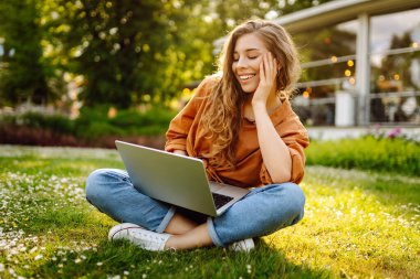 Yeşil çimlerde dizüstü bilgisayarla oturan mutlu kadın. Çevrimiçi eğitim, serbest çalışma, online alışveriş, blog yazma.