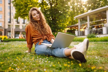 Yeşil çimlerde dizüstü bilgisayarla oturan mutlu kadın. Çevrimiçi eğitim, serbest çalışma, online alışveriş, blog yazma.