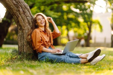 Yeşil çimlerde dizüstü bilgisayarla oturan mutlu kadın. Çevrimiçi eğitim, serbest çalışma, online alışveriş, blog yazma.