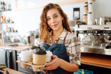 Paket yemek konsepti. Barda önlüklü neşeli bir kadın bir kafede kahve bardakları tutuyor. Yiyecek ve içecek..
