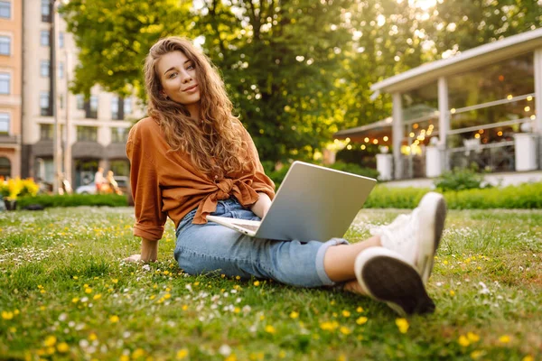 Yeşil çimlerde dizüstü bilgisayarla oturan mutlu kadın. Çevrimiçi eğitim, serbest çalışma, online alışveriş, blog yazma.