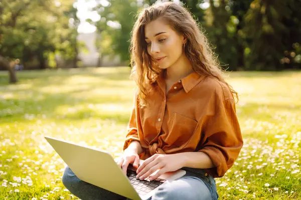 Yeşil çimlerde dizüstü bilgisayarla oturan mutlu kadın. Çevrimiçi eğitim, serbest çalışma, online alışveriş, blog yazma.