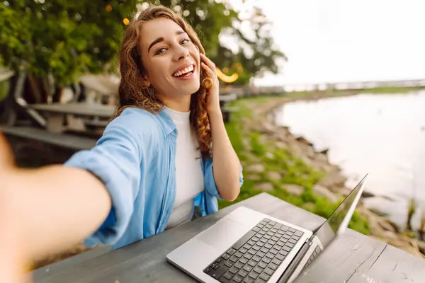 Selfie zamanı. Açık havada bir masada oturan mutlu bir kadın selfie çekiyor, göl manzarası güzel olan bir dizüstü bilgisayarla çalışıyor. Bloglama, serbest çalışma, online eğitim.