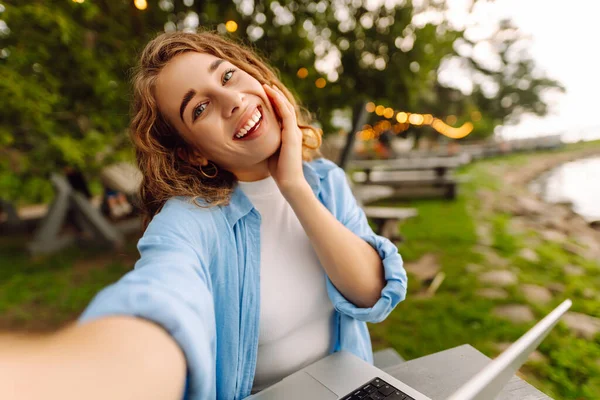 Selfie zamanı. Açık havada bir masada oturan mutlu bir kadın selfie çekiyor, göl manzarası güzel olan bir dizüstü bilgisayarla çalışıyor. Bloglama, serbest çalışma, online eğitim.