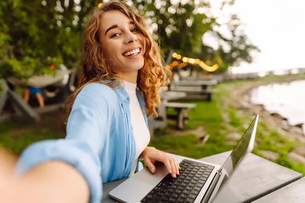 Selfie zamanı. Açık havada bir masada oturan mutlu bir kadın selfie çekiyor, göl manzarası güzel olan bir dizüstü bilgisayarla çalışıyor. Bloglama, serbest çalışma, online eğitim.