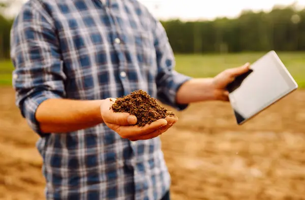 Farm hand holds black soil in her hands and checking with a digital tablet. Concept of agriculture, technology, business and ecology.