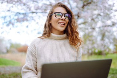 Parktaki dizüstü bilgisayarlı genç kıvırcık kadın. Gülümseyen serbest çalışan, dışarıda çalışan, güzel havanın tadını çıkaran biri. Eğitim, iş, blog veya serbest çalışma kavramı.