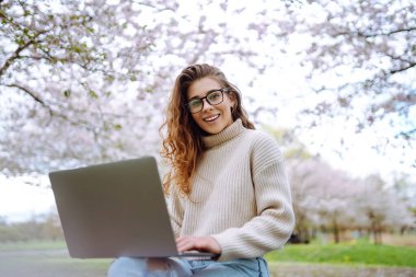 Parktaki dizüstü bilgisayarlı genç kıvırcık kadın. Gülümseyen serbest çalışan, dışarıda çalışan, güzel havanın tadını çıkaran biri. Eğitim, iş, blog veya serbest çalışma kavramı.