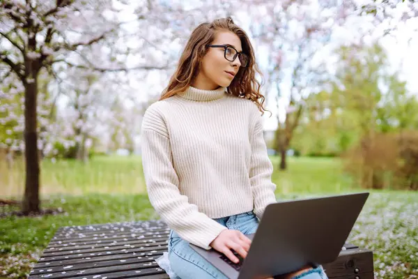 Parktaki dizüstü bilgisayarlı genç kıvırcık kadın. Gülümseyen serbest çalışan, dışarıda çalışan, güzel havanın tadını çıkaran biri. Eğitim, iş, blog veya serbest çalışma kavramı.