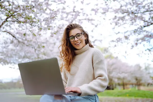 Parktaki dizüstü bilgisayarlı genç kıvırcık kadın. Gülümseyen serbest çalışan, dışarıda çalışan, güzel havanın tadını çıkaran biri. Eğitim, iş, blog veya serbest çalışma kavramı.