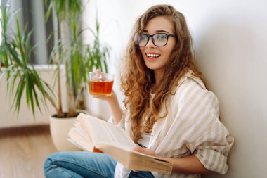 Genç bir kadın yerde oturuyor, çay içiyor ve kitap okuyor. Yaşam tarzı, rahatlama ve ev hayatı, konfor konsepti.