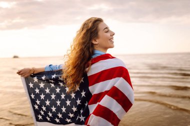 Amerika Birleşik Devletleri bayrağını tutan ve gün batımında sahilde koşan mutlu kadın. Vatansever bayram. ABD 4 Temmuz 'u kutluyor. Bağımsızlık Günü kavramı