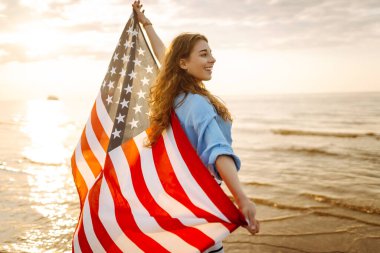 Amerika Birleşik Devletleri bayrağını tutan ve gün batımında sahilde koşan mutlu kadın. Vatansever bayram. ABD 4 Temmuz 'u kutluyor. Bağımsızlık Günü kavramı