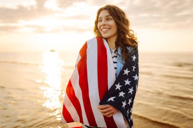 Amerika Birleşik Devletleri bayrağını tutan ve gün batımında sahilde koşan mutlu kadın. Vatansever bayram. ABD 4 Temmuz 'u kutluyor. Bağımsızlık Günü kavramı