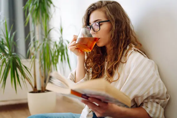 Young woman sitting on floor, drinking tea and reading book. Lifestyle, relaxation and domestic life, comfort concept.