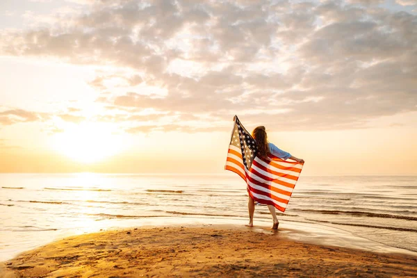 Amerika Birleşik Devletleri bayrağını tutan ve gün batımında sahilde koşan mutlu kadın. Vatansever bayram. ABD 4 Temmuz 'u kutluyor. Bağımsızlık Günü kavramı