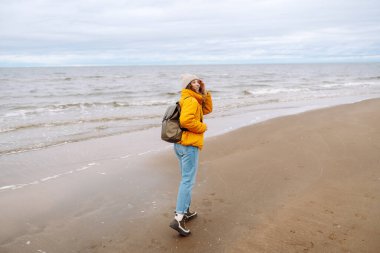 Sarı ceketli ve deniz kenarında poz veren mutlu bir turist. Seyahat, yaşam tarzı, macera.