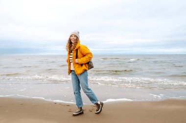 Sarı ceketli ve deniz kenarında poz veren mutlu bir turist. Seyahat, yaşam tarzı, macera.