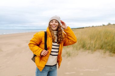 Sarı ceketli ve deniz kenarında poz veren mutlu bir turist. Seyahat, yaşam tarzı, macera.