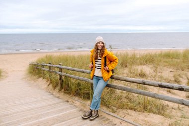Sarı ceketli ve deniz kenarında poz veren mutlu bir turist. Seyahat, yaşam tarzı, macera.