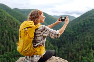 Turist kadın hatıralar için açık havada fotoğraf çekiyor, vadi dağları manzaralı uçurumun tepesinde selfie çekiyor, seyahat maceralarını paylaşıyor.. 