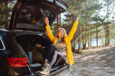 Genç bir kadın ormanda bir arabanın bagajında oturuyor. Arabayla seyahat etme kavramı, aktif yaşam tarzı..