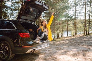 Genç bir kadın ormanda bir arabanın bagajında oturuyor. Arabayla seyahat etme kavramı, aktif yaşam tarzı..