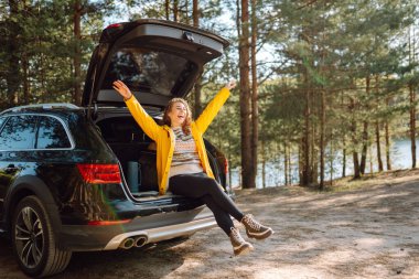 Genç bir kadın ormanda bir arabanın bagajında oturuyor. Arabayla seyahat etme kavramı, aktif yaşam tarzı..