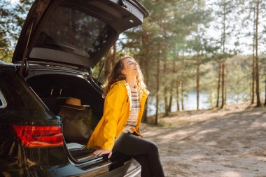 Genç bir kadın ormanda bir arabanın bagajında oturuyor. Arabayla seyahat etme kavramı, aktif yaşam tarzı..