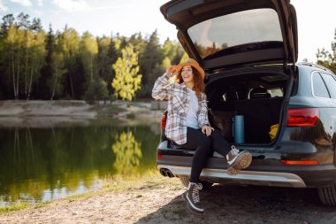 Genç bir kadın ormanda bir arabanın bagajında oturuyor. Arabayla seyahat etme kavramı, aktif yaşam tarzı..