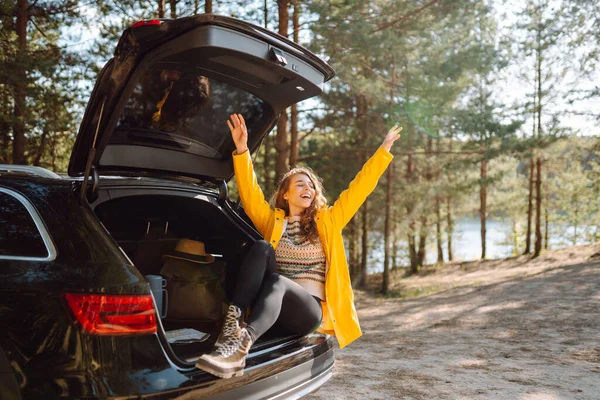 Genç bir kadın ormanda bir arabanın bagajında oturuyor. Arabayla seyahat etme kavramı, aktif yaşam tarzı..