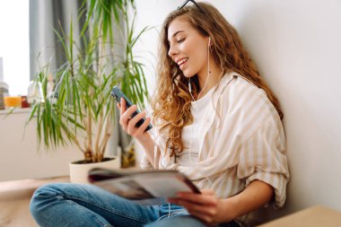 Genç kadın evde oturuyor, akıllı telefon kullanıyor, öğreniyor, kitap okuyor. Dinlenme, eğitim, teknoloji kavramı.