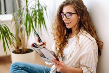 Genç kadın evde oturuyor, akıllı telefon kullanıyor, öğreniyor, kitap okuyor. Dinlenme, eğitim, teknoloji kavramı.