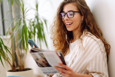 Genç kadın evde oturuyor, akıllı telefon kullanıyor, öğreniyor, kitap okuyor. Dinlenme, eğitim, teknoloji kavramı.