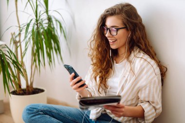 Genç kadın evde oturuyor, akıllı telefon kullanıyor, öğreniyor, kitap okuyor. Dinlenme, eğitim, teknoloji kavramı.