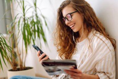 Genç kadın evde oturuyor, akıllı telefon kullanıyor, öğreniyor, kitap okuyor. Dinlenme, eğitim, teknoloji kavramı.