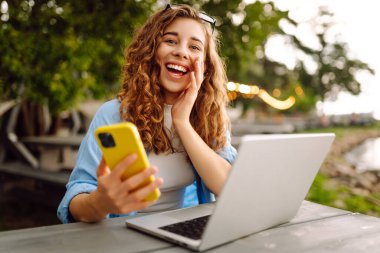 Diz üstü bilgisayarı ve telefonu olan genç bir kadın gölün yakınındaki açık bir kafede bir masada. Eğitim çevrimiçi. IT, uzaktan kumanda. İş, blog yazma, serbest çalışma kavramı.