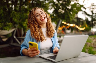 Diz üstü bilgisayarı ve telefonu olan genç bir kadın gölün yakınındaki açık bir kafede bir masada. Eğitim çevrimiçi. IT, uzaktan kumanda. İş, blog yazma, serbest çalışma kavramı.