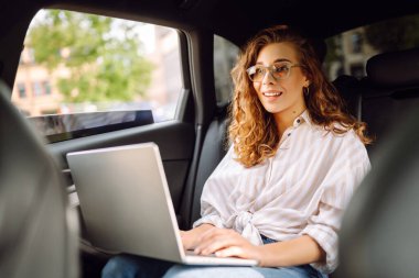 Bir arabanın arka koltuğunda dizüstü bilgisayarı olan genç bir kadın. Eğitim çevrimiçi. Uzaktan kumanda. İş, blog yazma, serbest çalışma kavramı.