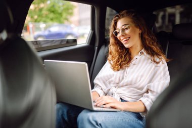 Bir arabanın arka koltuğunda dizüstü bilgisayarı olan genç bir kadın. Eğitim çevrimiçi. Uzaktan kumanda. İş, blog yazma, serbest çalışma kavramı.