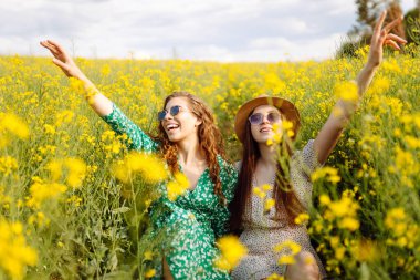 Gülümseyen kadın turistler çiçek tarlasında yürürler, sarı çiçeklere dokunurlar. Doğa, tatil, rahatlama ve yaşam tarzı. Yaz manzarası.