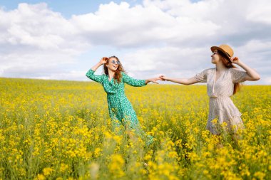 Gülümseyen kadın turistler çiçek tarlasında yürürler, sarı çiçeklere dokunurlar. Doğa, tatil, rahatlama ve yaşam tarzı. Yaz manzarası.