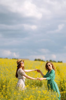 Gülümseyen kadın turistler çiçek tarlasında yürürler, sarı çiçeklere dokunurlar. Doğa, tatil, rahatlama ve yaşam tarzı. Yaz manzarası.