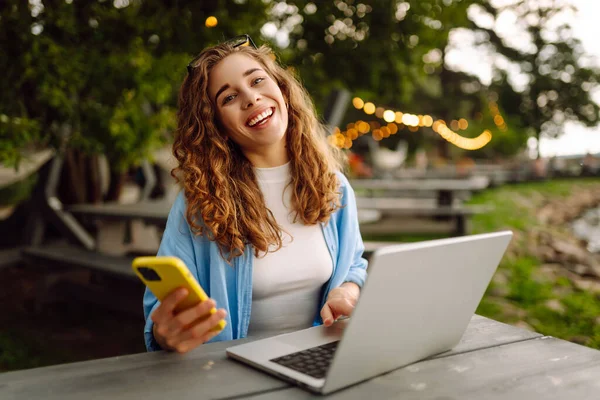 Diz üstü bilgisayarı ve telefonu olan genç bir kadın gölün yakınındaki açık bir kafede bir masada. Eğitim çevrimiçi. IT, uzaktan kumanda. İş, blog yazma, serbest çalışma kavramı.