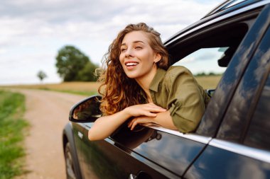 Arabanın camından sarkıp doğanın tadını çıkaran genç bir kadın. Kız dinleniyor ve yolculuğun tadını çıkarıyor. Yaşam tarzı, seyahat, turizm, aktif yaşam.