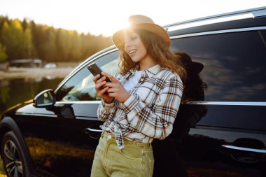 Gün batımında akıllı telefon kullanan genç ve güzel bir kadın arabayla seyahat ediyor. Boş zaman, seyahat, teknoloji, blog.