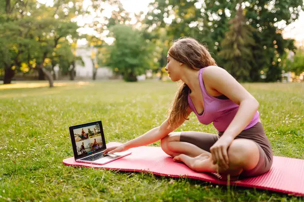 Laptopun önünde yoga yapan genç bir kadın, parkta online dersler izliyor. Antrenman egzersizi. Sağlıklı yaşam tarzı, spor. Çevrimiçi eğitim..