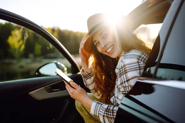 Young beautiful woman traveling by car using smartphone at sunset. Leisure, travel, technology, blogging.