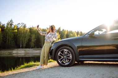 Tatilde mutlu bir kadın arabasının yanında akıllı telefonuyla selfie çekiyor. Araba yolculuğunda şık bir model. Seyahat, günlük tutma kavramı.